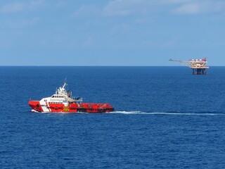 Red supply vessel navigates the deep blue ocean, heading towards an offshore oil platform under a clear sky. A captivating scene of maritime industry and vastness.