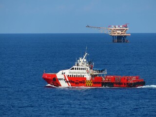 Red supply vessel navigates the deep blue ocean, approaching a distant offshore oil platform under a clear sky. A scene of maritime industry and vastness.