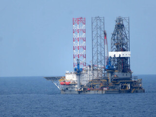 A large offshore oil rig, composed of multiple metal structures, floats on a calm blue ocean under a clear sky. The rig is the central focus, with the ocean forming a solid background. 