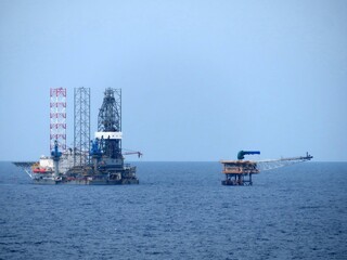 Two offshore oil rigs on a calm blue sea under a light blue sky. One rig is larger, more complex. The background is a clear, solid sky and sea. The scene is tranquil yet industrial.
