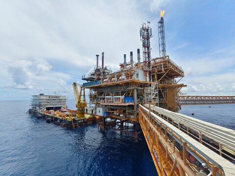 Vast offshore oil platform, a complex network of structures and pipelines, dominates the deep blue sea under a partly cloudy sky. A support vessel is nearby.