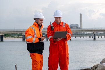 Two workers in orange overalls discuss project details near a river under a cloudy sky