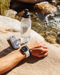 Wearable hydration monitor on a person's wrist, placed beside a transparent water bottle and smart health device on a sunlit rock near a clear stream — ai generative.