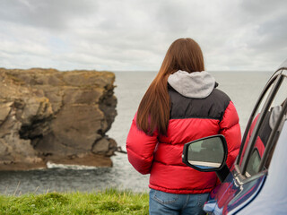 A woman in a red jacket stands next to a blue car in a parking lot and looking at a stunning nature scene with cliff, rough stone coastline and cloudy sky. Travel and tourism. West of Ireland.