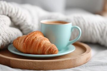A fresh croissant and a cup of coffee presented on a wooden tray, capturing the essence of a delightful morning breakfast in a cozy setting, with a soft blanket in the background.