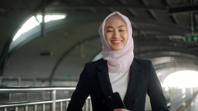 Happy asian muslim business woman wearing hijab holding and using smartphone waiting railway or skytrain at train station. Islamic woman people lifestyle travel. Muslim people concept.
