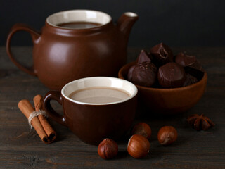 A wooden table with a jug and a cup of coffee along with a few hazelnuts, chocolate pralines, and cinnamon sticks evoking a sense of comfort and simplicity.
