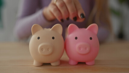 Woman saving money with two piggy banks on a wooden table in a cozy living room setting, highlighting ideas of financial planning and personal finance at home.