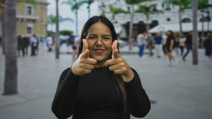 Fototapeta premium Smiling woman points fingers with confidence on a bustling street with people strolling by during a sunny day.
