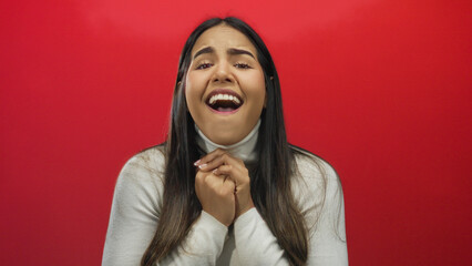 Woman expressing emotion with hands clasped, standing against a vibrant red background, showcasing...
