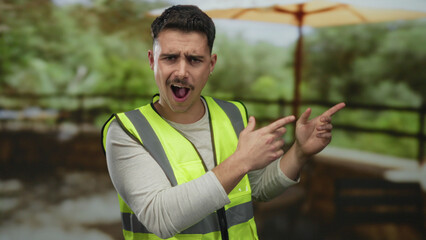 Young man in reflective vest points surprised in a park setting with lush greenery under a large...