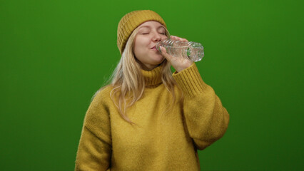 Young woman wearing yellow sweater and hat drinks water against a vibrant green background...
