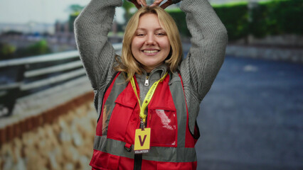 Volunteer woman with blonde hair making heart symbol on city street, wearing red vest and smiling...