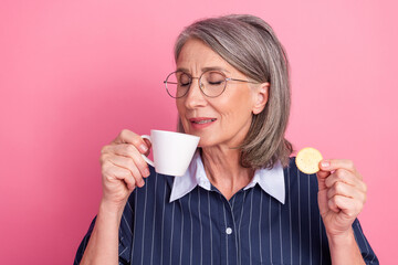 Mature woman with glasses sipping coffee and holding biscuit against a pink background while...