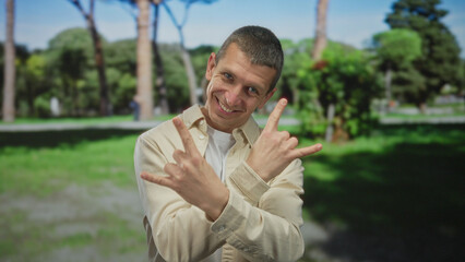 Caucasian man enthusiastically gestures rock hands in a sunny park, smiling amid lush green outdoor...