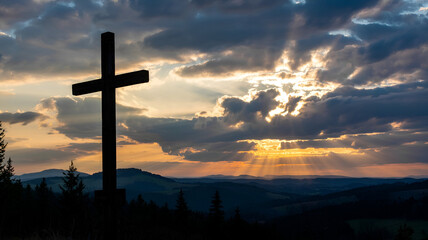 Silhouette of a cross against a dramatic sunset sky over a mountain range.