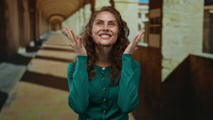 Woman smiling in a university hallway showcasing joyful emotions and expressive gestures while wearing a green sweater in an old academic setting with brick details.