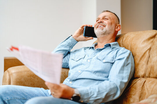 Man smiles while talking on the phone and reviewing documents in a cozy living room setting during daytime