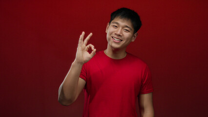 Naklejka premium Young chinese man in red shirt shows okay sign against isolated red background conveying approval and confidence