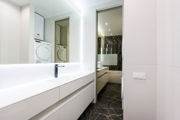 modern bathroom with white cabinets, a sleek sink, and a large mirror reflecting laundry machines. A bathroom is visible through a doorway, featuring dark marble