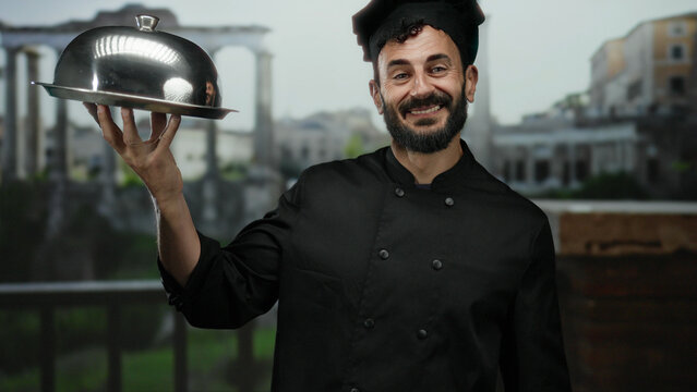 Chef with beard in black uniform holds a silver platter smiling in old town rome with ancient architecture in the background, showcasing culinary artistry.