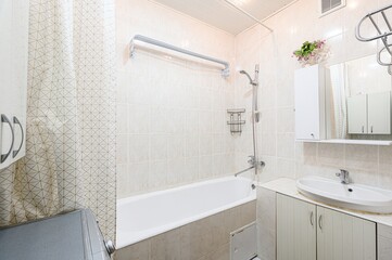 bright bathroom features beige tiles, a white bathtub, and a sink with cabinets. A shower curtain...