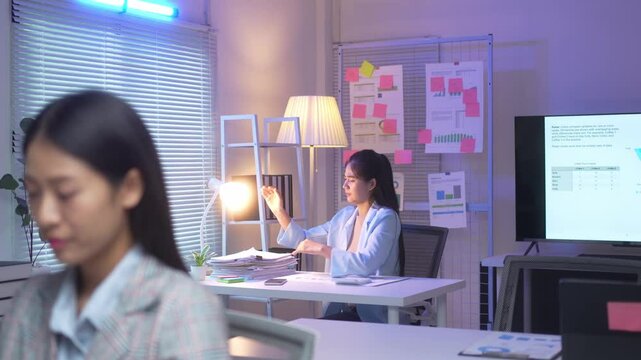 A woman is sitting at a desk with a stack of papers in front of her. She is wearing a blue shirt and she is focused on her work. The room is well-lit and has a modern, professional feel