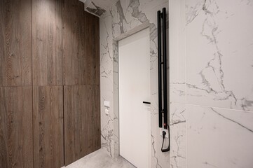 modern interior view featuring a wooden cabinet, a white door set against marble-patterned tiles, and a black towel warmer. The flooring appears to be a light gray