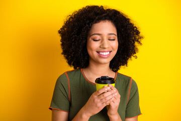 Young woman smiling happily in casual attire, holding a cup, standing against a vibrant yellow...