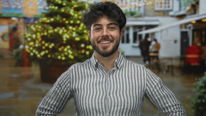 Fototapeta premium Young hispanic man with beard smiling outdoors on a street with festive lights and a christmas tree, wearing a striped shirt and casual attire, enjoying the holiday atmosphere.