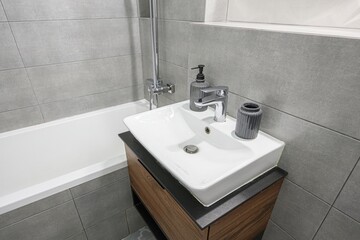 Modern bathroom sink with a white basin, wooden cabinet, gray tiles, and chrome fixtures. A soap dispenser and decorative vase adorn the countertop