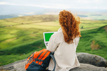 Person works on a laptop while sitting on a rock overlooking green hills on a cloudy day in a rural landscape