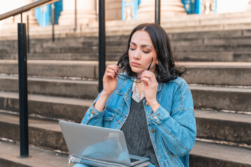 Fototapeta premium Young woman working on laptop while sitting on steps of historic building in city during bright day