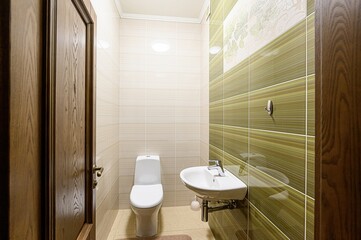 small, modern bathroom with beige and green tiles, featuring a toilet, sink, and wooden door. Clean and simple design
