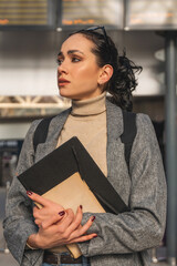 Businesswoman in a gray blazer holding a folder while looking thoughtful in a bustling urban...