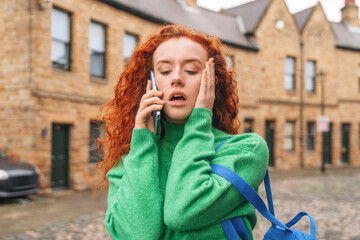 Red-haired woman standing on a cobblestone street talking on her phone while showing signs of distress in a historic neighborhood