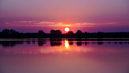 Naklejka premium Serene sunset over a calm lake with silhouetted trees