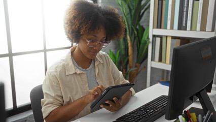 Woman with curly hair at her office desk using a tablet, surrounded by indoor plants and bookshelves, epitomizing a modern workplace environment with natural light.