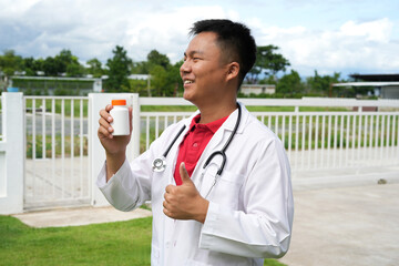 Doctor with stethoscope who is standing in Hospital garden is holding an medicine bottle to assess...