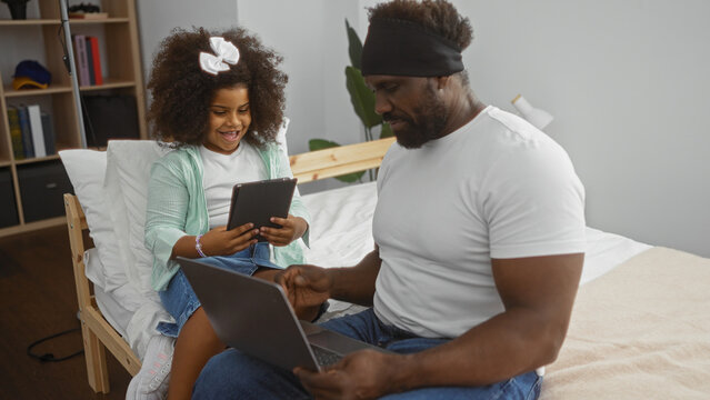 Father and daughter sitting on a bed in a cozy room using laptop and tablet, showcasing family bond and digital lifestyle at home.
