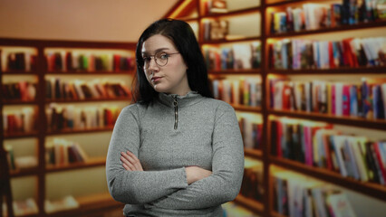 Woman with glasses and brunette hair sits indoors in a library with bookshelves, wearing a grey...