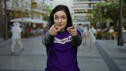Woman pointing confidently at camera wearing volunteer shirt on bustling street with background of...