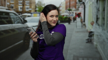 Woman with brunette hair smiling while holding a smartphone on a street in a volunteer shirt outdoors, surrounded by blurred vehicles and buildings in an urban area.