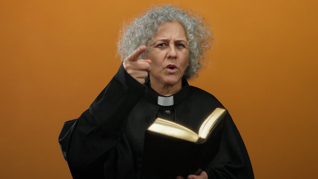 Elderly woman with grey hair wearing clergy attire reads from a book, pointing emphatically, set against an isolated orange background wall.