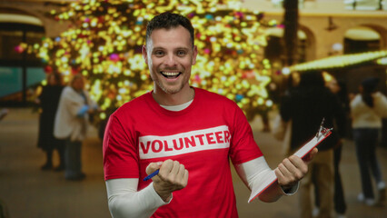 Man in red volunteer shirt smiles holding clipboard with holiday lights in urban area street in...