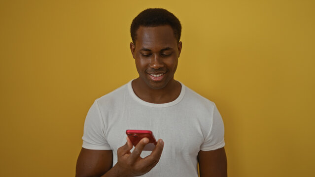 Young man smiling while looking at smartphone over vibrant yellow background, conveying modern lifestyle and connectivity in an isolated setting.