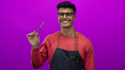 Young hispanic man holds hairdressing scissors against a vibrant pink background, wearing glasses,...