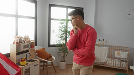 Young man in pink sweater thoughtfully standing in modern nursery room with crib, soft toys, and bright natural lighting, conveying a serene and cozy domestic atmosphere.