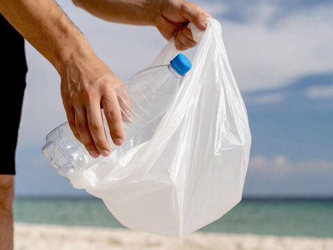 On the beach, a man holds a plastic bag into which he is collecting plastic bottles from the shore, and around him are sand and waves. World Environment Day.