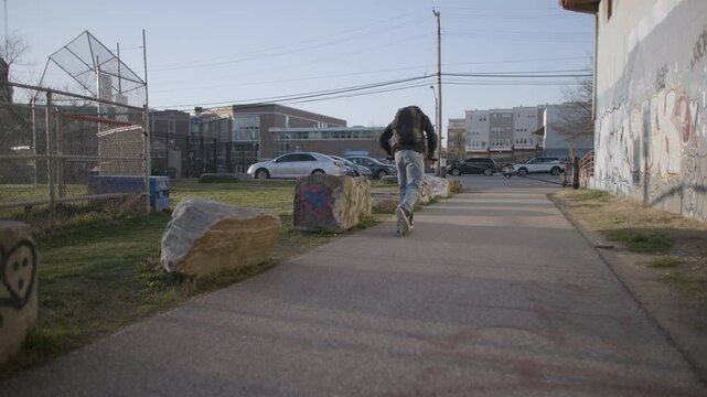 Boy rides scooter on a paved road towards a school, passing a graffitied wall.